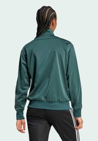 Green satin zip-up jacket with a high collar, ribbed cuffs, and waistband, worn over black pants featuring white side stripes.