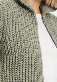 Stickad cardigan i ljusgrön. Har ett texturerat mönster, öppen framsida och krage. Bärs över en vit skjorta. Mjuk, fyllig garn.