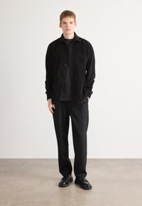 Young man standing indoors wearing a black corduroy shirt over a dark t-shirt, black trousers, and black leather shoes.