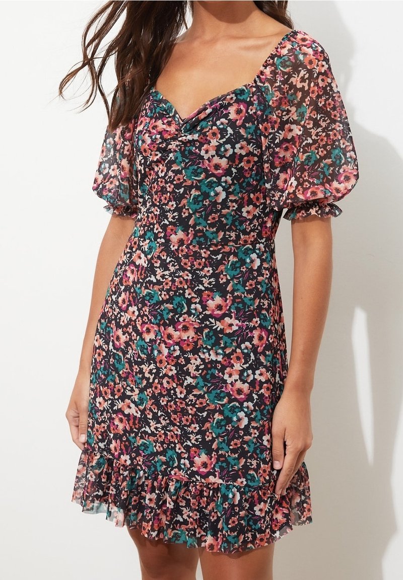 Femme portant une robe noire à fleurs avec des manches bouffantes et un ourlet volanté, featuring des fleurs roses, bleu sarcelle et blanches sur un fond uni.