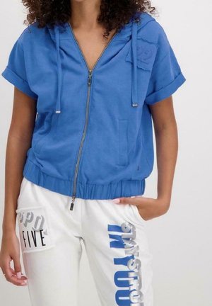 Woman wearing a blue short-sleeve zip-up hoodie and white pants with large blue and black text on the front.