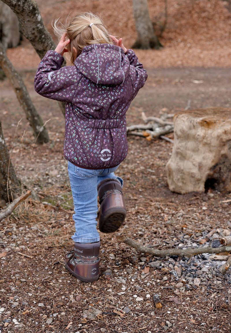 Paarse jas met bloemenpatroon, elastiek in de taille, gecombineerd met een blauwe spijkerbroek en bruine, geblokte laarzen, tegen de achtergrond van een bosgrond.