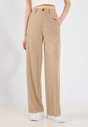 Beige bukser med højtaljede og vide ben, båret med hvide sneakers, den ene hånd i lommen, vist fra taljen til fødderne på en ensfarvet baggrund.