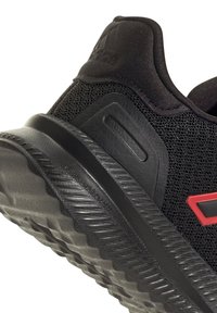 Zapatilla deportiva negra con parte superior de malla, suela texturizada y acentos rojos. El diseño incluye un cuello acolchado y detalles de refuerzo resistentes.