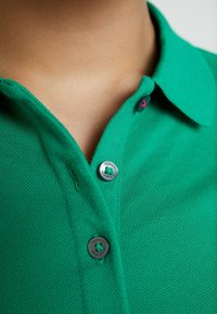 Zielona koszulka polo z teksturowanej tkaniny, z dwoma guzikami z metalowym logotypem 'Tommy Hilfiger' i kontrastowym różowym akcentem na górnym guziku.