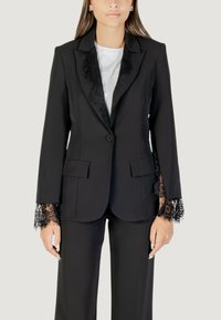Blazer nero con rifiniture in pizzo sui polsini e sulle rever, caratterizzato da una chiusura a singolo bottone e due tasche frontali, abbinato a pantaloni coordinati.