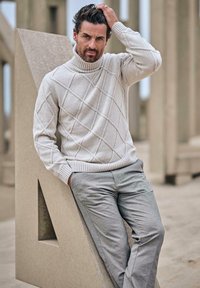 Hellgrauer, gestrickter Rollkragenpullover mit Rautenmuster; gerippte Bündchen und Saum. Kombiniert mit hellgrauen Hosen. Strukturierter Hintergrund.