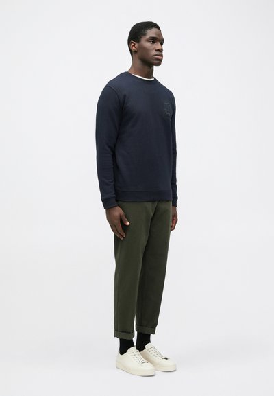 Marineblå sweatshirt med rund hals og broderede detaljer, parret med olivengrønne tapered bukser og hvide sko. Enkel, afslappet design.