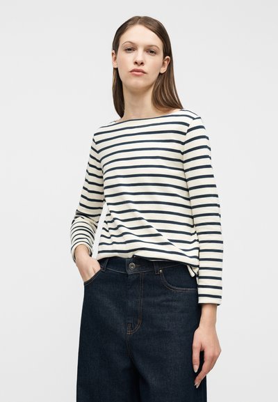 Γυναίκα που φοράει λευκό μπλουζάκι με μαύρες οριζόντιες ρίγες και σκούρο μπλε τζιν ψηλόμεσο, στέκεται με το ένα χέρι στην τσέπη.