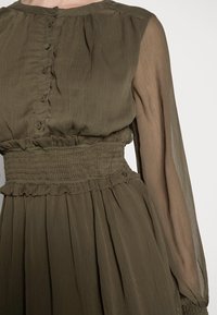 Vestido verde oliva con mangas largas translúcidas, cintura fruncida y detalles de botones en la parte delantera. Tejido ligero y texturizado con una falda fluida.