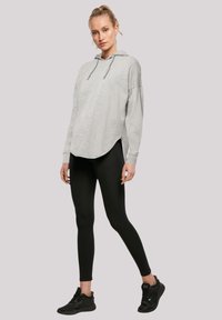 Hellgrauer Hoodie mit abgerundetem Saum und seitlichen Schlitzen, kombiniert mit schwarzen Leggings und schwarzen Sportschuhen. Glatte Textur, lässiger Schnitt.
