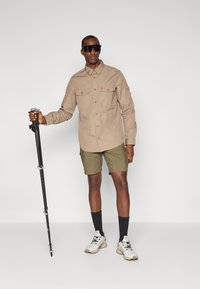 Camicetta button-up marrone chiaro con due tasche sul petto, pantaloni corti verdi, bastoni da trekking, scarpe bianche e beige, e occhiali da sole scuri. Sfondo neutro.