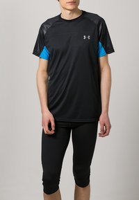 Camiseta deportiva negra con acentos azules y un diseño texturizado; combinada con leggings ajustados de longitud capri en negro. Presenta un logo en el pecho.