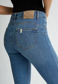 Jeans de mezclilla azul de tiro alto que presentan una etiqueta de marca texturizada en color marrón, un pequeño bolsillo trasero y detalles de costura visibles en un acabado suave.