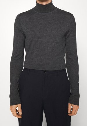Personne portant un pull à col roulé gris foncé ajusté, rentré dans un pantalon noir taille haute, debout devant un fond uni.