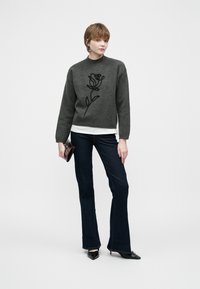 Mulher a usar um suéter cinza escuro com um contorno de rosa preta, jeans flare escuros, saltos agudos pretos, segurando uma pequena carteira escura.