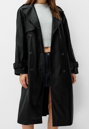 Zwarte dubbelgeknoopte lange trenchcoat met knoopmanchetten gedragen over een witte geribde cropped trui en een donkere denim rok.