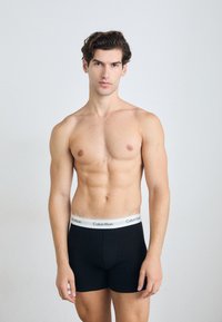 Fekete pamut boxer alsónadrág, fehér Calvin Klein logós derékpánttal. A modellen nincs póló, jól láthatóan izmos testalkattal és egyenes tartással rendelkezik.
