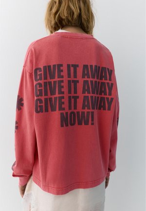 Person with shoulder-length hair wears a red sweatshirt with bold black text "GIVE IT AWAY GIVE IT AWAY GIVE IT AWAY NOW!" on the back.