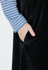 Schwarzes Cordkleidungsstück mit sichtbarem Taschen, kombiniert mit einem blau-weiß gestreiften Langarmhemd. Texturierte, gerippte Stoffdetails.