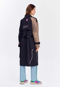 Čierny trenčkot s rukávmi z leopardieho vzoru, s viazaním v páse a vyšívaným textom. Vyznačuje sa kontrastnými textúrami a farbami.