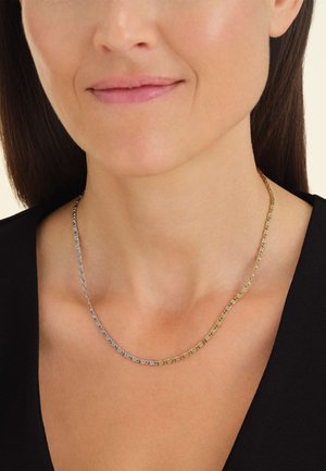 Dunne halsketting met afwisselende zilveren en gouden schakels in een gebogen ontwerp, rustend op het sleutelbeen, gedragen over een zwart top.