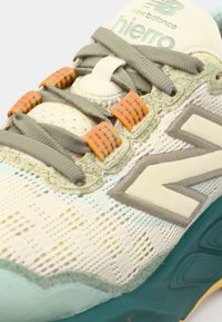 La chaussure présente un dessus en mesh respirant crème avec des accents vert menthe, des tons terreux et des lacets orange. Détails texturés notables et logo.