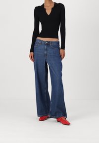 Černý žebrovaný top s dlouhým rukávem a výstřihem do V, spárovaný s modrými džíny s širokými nohavicemi a červenými plochými botami. Jednoduchý, neformální outfit s texturovanými materiály.