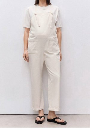 Beige overalls van katoen met verstelbare bandjes, voorzakken en een losse pasvorm. Draagt over een wit T-shirt en gecombineerd met zwarte slippers.