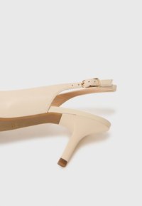 Scarpa con tacco alto slingback beige in pelle liscia con dettaglio della fibbia dorata, caratterizzata da un design elegante e un tacco affusolato.