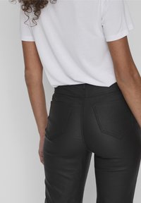 Pantalones negros de cintura alta con un acabado texturizado, que cuentan con dos bolsillos traseros, detalles cosidos y un corte clásico, combinados con una camiseta blanca.