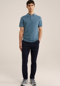 Polo de color verde azulado con rayas verticales sutiles, mangas cortas y un frente con tres botones, combinado con pantalones azul marino oscuros y zapatillas grises.