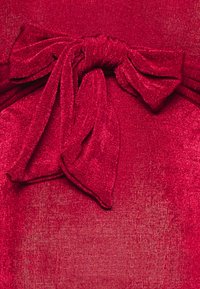 Tecido vermelho com uma textura cintilante, apresentando um detalhe de laço proeminente feito do mesmo material, criando um design coeso.