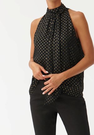 Vrouw draagt een mouwloos zwart haltertopje met klein goudkleurig cirkelpatroon, gecombineerd met zwarte broek, handen in de taille geklemd.