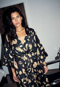 Čierny plášť zdobený zlatými motívmi, so špicatým výstrihom do V, širokými rukávmi a opaskom viazaným v páse. Hladká textúra, moderný dizajn.