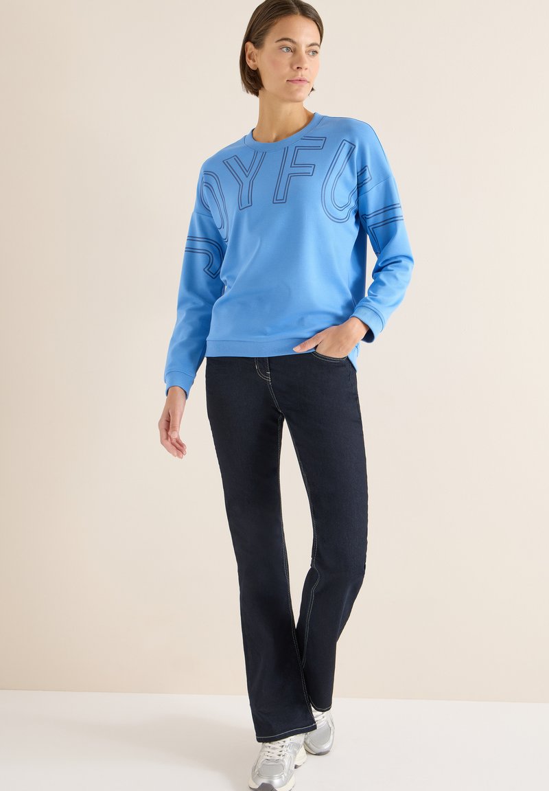 Jasnoniebieska bluza z napisem „JOYFUL”, zestawiona z ciemnymi dzwonowatymi spodniami dżinsowymi i srebrnymi butami sportowymi. Gładka tkanina, casualowy design.