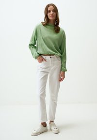 Maglietta a maniche lunghe verde a coste, con collo alto, abbinata a jeans bianchi a gamba dritta e sneakers beige, in piedi davanti a uno sfondo neutro.