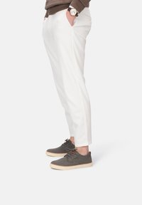 Mann in weißen schmalen Hosen, grauen lässigen Schnürschuhen, braunem Pullover und mit einer Armbanduhr, die die Hände in den Taschen hat.