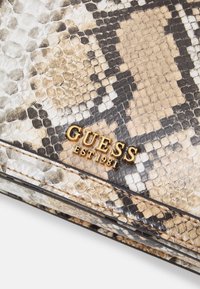Portafoglio con motivo pelle di serpente in beige, nero e bianco. Presenta un logo metallico dorato "GUESS EST 1981" e dettagli con cuciture nere.