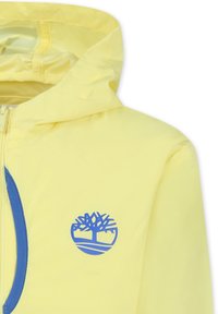 Giacca impermeabile gialla con zip frontale e logo blu. Presenta un cappuccio e accenti blu a contrasto lungo la zip.