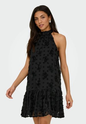 Femme portant une robe noire sans manches avec des motifs floraux en 3D et un ourlet volanté, debout devant un fond clair uni.