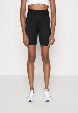 Kobiecy tułów i nogi w czarnych, sportowych szortach z wysokim stanem z białym logo Nike, białym staniku sportowym i białych sneakersach.
