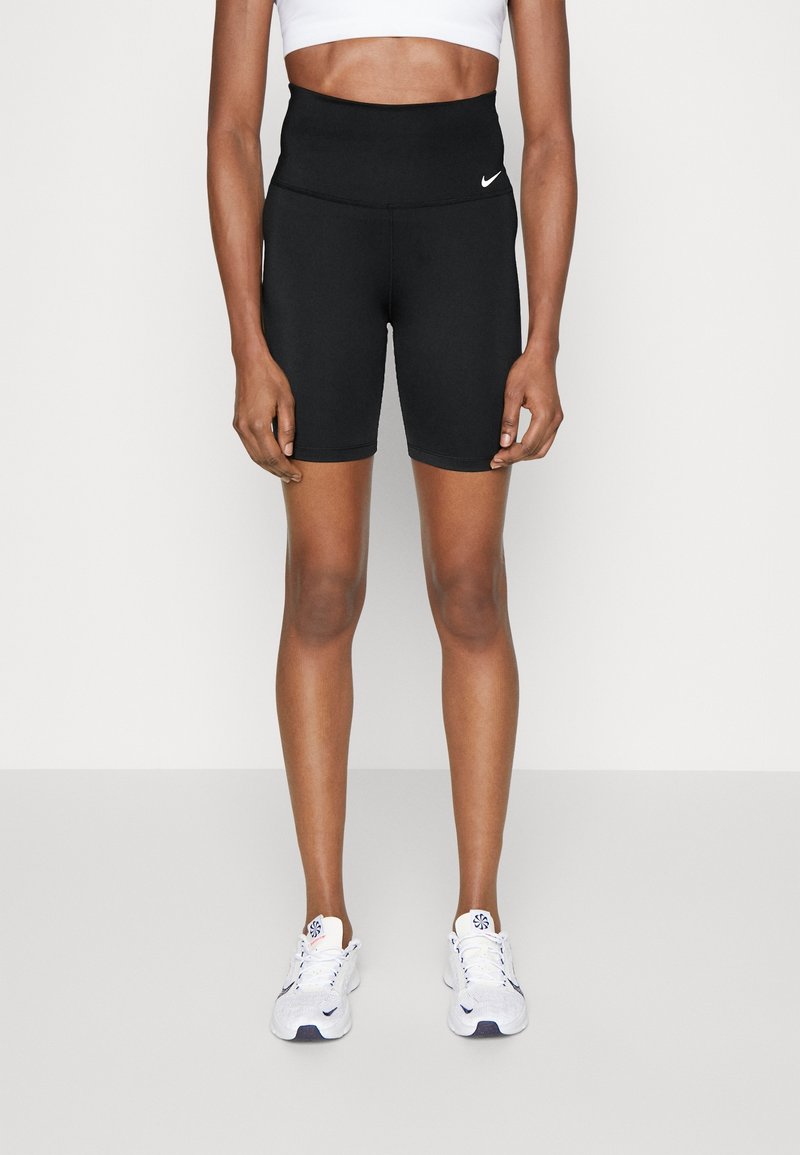 Buste et jambes féminins portant un short de sport noir taille haute avec un logo Nike blanc, un soutien-gorge de sport blanc et des baskets blanches.
