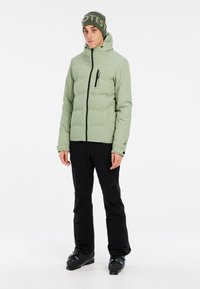 Hellgrüne Pufferjacke mit Kapuze, Front-Reißverschluss und Reißverschluss-Tasche, kombiniert mit schwarzen Hosen und Skischuhen, einfaches Design, glatte Textur.