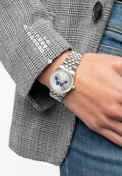 Strieborné hodinky s motýlím dizajnom na ciferníku, nosené na zápästí so strieborným kovovým náramkom, v kombinácii so sivým károvaným sako.