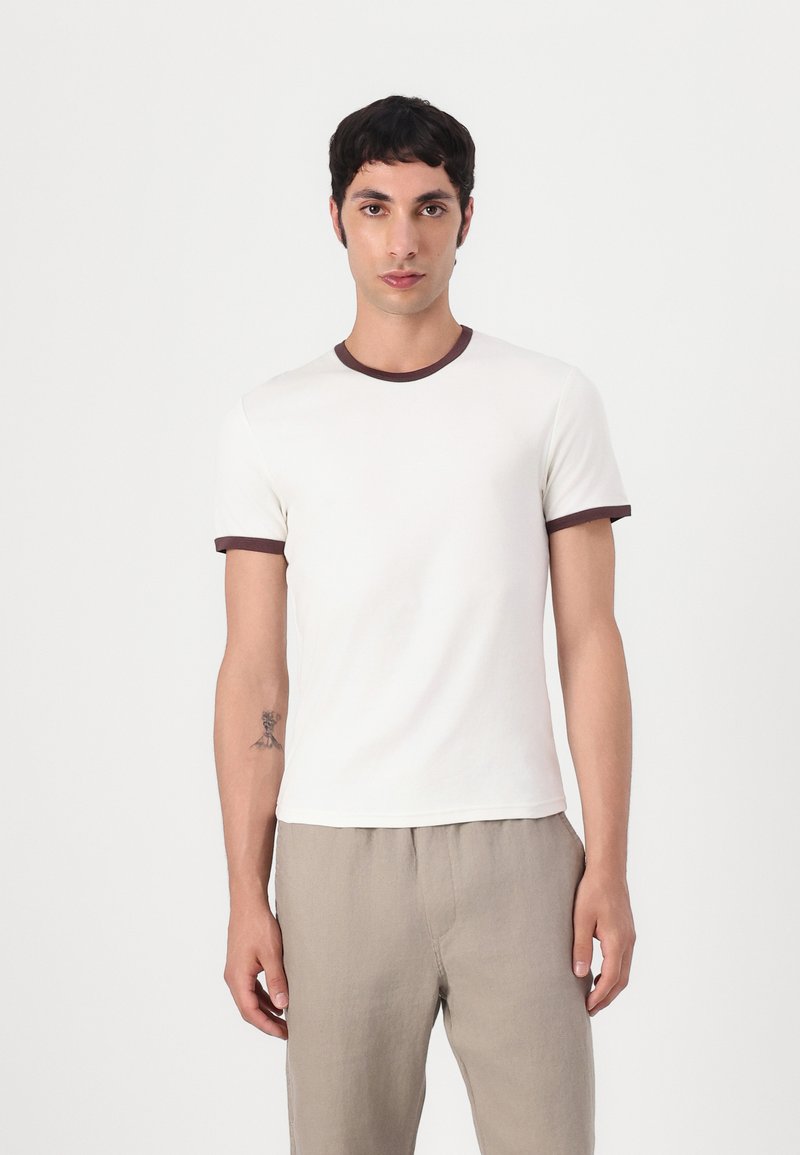 White cotton t-shirt with brown ribbed neckline and sleeves, featuring a fitted design. The model wears beige trousers and displays a tattoo on the arm.