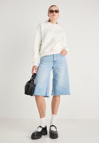 Felpa bianca con tasca frontale, pantaloni bermuda in denim azzurro chiaro con orli grezzi, scarpe nere chunky, borsa nera e calze bianche con bordi arricciati.