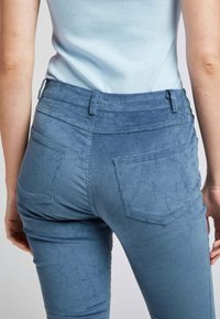 Pantalons en velours côtelé bleu clair avec une coupe slim. Présentent des côtes verticales, deux poches arrière et une taille haute. La texture semble douce et veloutée.