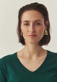Pull vert à col en V en matériau doux ; avec une texture subtile. De grandes boucles d'oreilles dorées mettent en valeur le décolleté.