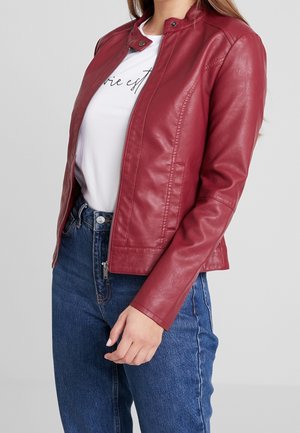 Woman wearing a red leather jacket over a white printed t-shirt and blue denim jeans, standing against a plain gray background.